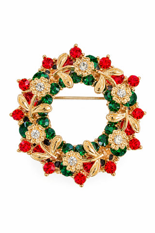 A gold-tone floral wreath brooch decorated with red and green faceted rhinestones, resting on a textured dark gray surface.